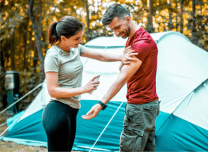 The campsite is all set, the tent is up. A women is spraying minus bite all natural bug spray on a man.
